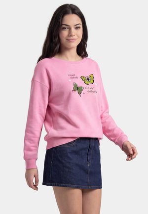 Giovane donna che indossa una felpa rosa con stampe di farfalle colorate e una gonna di jeans blu scuro, in piedi davanti a uno sfondo chiaro e uniforme.