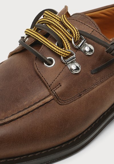Chaussure en cuir marron avec des lacets rayés jaunes et marron passés dans des œillets métalliques et des crochets argentés, montrant des coutures détaillées et une texture.