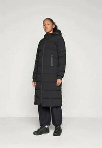 Mladý človek stojí oblečený v dlhom čiernom kabáte s kapucňou, vo voľných čiernych nohaviciach a čiernych teniskách na jednoduchom svetlom pozadí.