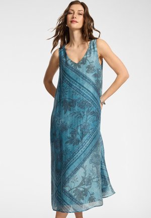 Frau trägt ärmelloses, blaues Maxikleid mit Blumenmuster und V-Ausschnitt, steht mit den Händen hinter dem Rücken vor schlichtem Hintergrund.