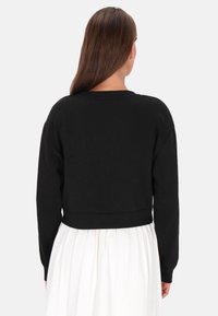 Sudadera negra recortada con mangas largas y dobladillo acanalado, mostrada en un modelo que lleva una falda blanca, con una textura suave.