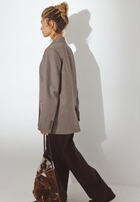 Übergroßer grauer Blazer mit glatter Textur, einreihiger Front und Manschetten mit vier Knöpfen, kombiniert mit schwarzen weit geschnittenen Hosen und einer braunen Handtasche mit Muster.