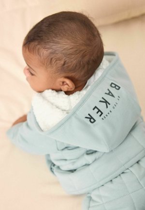 Niemowlę w jasnoniebieskiej kurtce z kapturem wyściełanym białym polarem, leżące na beżowej powierzchni, z głową odwróconą na bok.