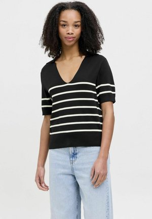 Suéter negro de cuello en V con mangas cortas que presenta rayas horizontales blancas. Tejido de punto suave con un corte relajado. Llevado con pantalones vaqueros de color azul claro.