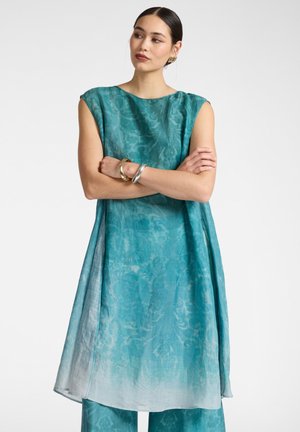 Femme aux cheveux foncés en chignon bas, portant une robe sans manches bleu sarcelle et un pantalon assorti, bras croisés, accessoirisée de bracelets dorés et argentés ainsi que des boucles d'oreilles.