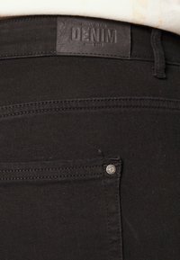 Os jeans de denim preto apresentam um tecido texturizado, um rótulo de couro com a descrição "DENIM" e um botão prateado visível perto do bolso.