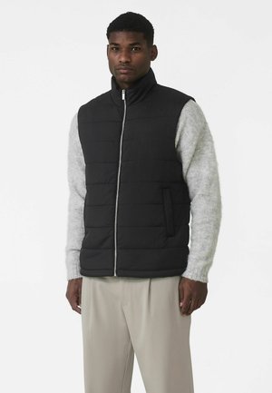 Czarna pikowana kamizelka z wysokim kołnierzem, zapinana na zamek błyskawiczny, z poziomym wzorem pikowania, noszona na jasno szarym swetrze. Spodnie są beżowe.