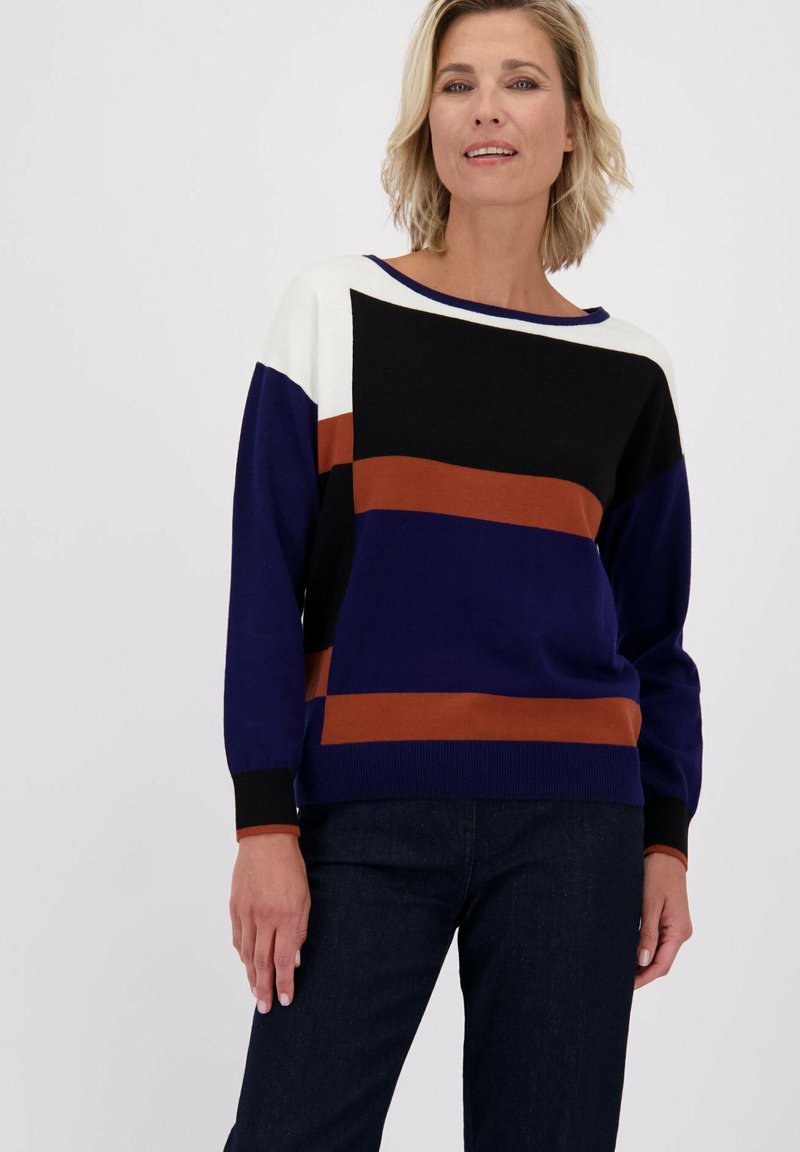 Blonde vrouw in een trui met lange mouwen, met kleurblokken in zwart, bruin, marineblauw en wit, gecombineerd met donkerblauwe jeans.