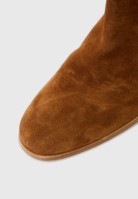 Bottine en suède marron avec un bout arrondi et une texture lisse, présentant une forme courbée et des coutures subtiles le long de la semelle.