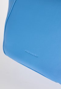 Borsa in eco-pelle blu con una texture liscia, forma arrotondata e logo in rilievo. Caratterizzata da un design minimalista e bordi puliti.