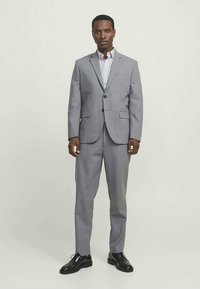 Homem vestindo um fato cinza claro de dois botões com calças a condizer, camisa riscada e sapatos pretos de gala, de pé contra um fundo simples.