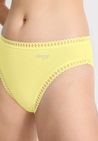 Close-up van een persoon die gele katoenen ondergoed draagt met een golvende kantafwerking en een klein 'sloggi'-logo aan de voorkant.