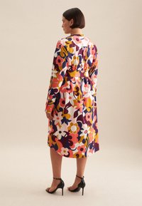 Kleid mit blumigem Muster in lebhaften Farben, mit langen Ärmeln, tailliertem Gürtel und knielangem Design. Getragen mit schwarzen High-Heels.