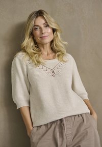 Lys beige strikket sweater med ribbet struktur, der har et blondermønster på skulderen og korte ærmer, parret med løse taupe bukser.