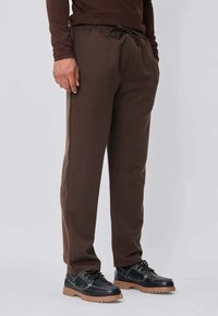 Personne portant un pantalon à cordon brun et des chaussures noires à lacets avec des semelles épaisses de couleur beige, debout devant un fond clair uni.