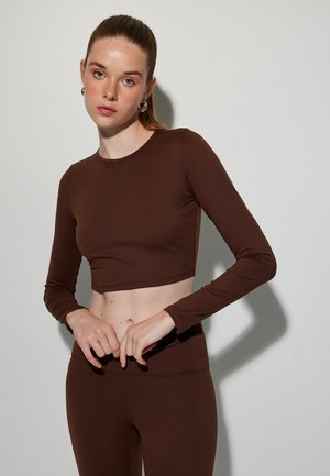 Brauner, cropped Langarmoberteil aus glattem Stoff, mit Rundhalsausschnitt und taillierter Silhouette. Getragen mit passenden Leggings.