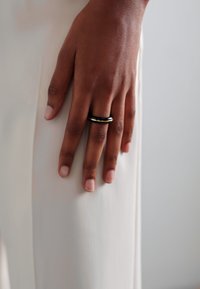Zlato-črna prstanska, ki jo nosi roka, naslonjena na svetlo obarvano tkanino, prikazuje gladko teksturo in preprost dizajn.