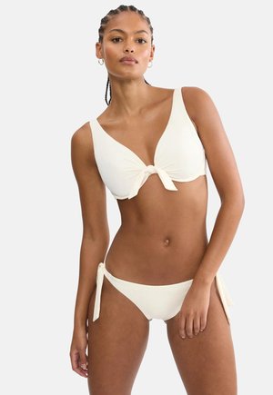 Kobieta w białym bikini z wiązanym na supeł topem i bokserkami z wiązaniem po bokach, stojąca na tle jasnego, jednolitego tła.