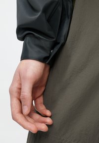 Temno siva jakna z teksturirano tkanino, z ozkim manšetom in kontrastnim olivno zelenim oblačilom spodaj. Roka počiva v bližini.