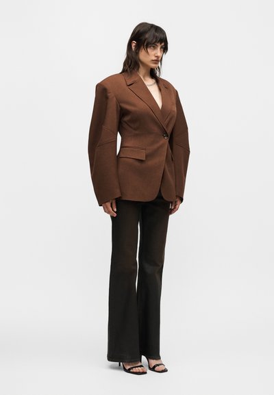 Γυναίκα που φορά καφέ oversized σακάκι με υπερβολικούς ώμους, μαύρο φαρδύ παντελόνι και μαύρες ανοιχτές γόβες, όρθια μπροστά σε απλό φόντο.