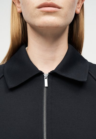 Γυναίκα με ανοικτόχρωμο δέρμα και ίσια ξανθά μαλλιά φοράει μαύρο σακάκι με γιακά που έχει φερμουάρ κλειστό μέχρι τον λαιμό, εμφανίζεται από το πηγούνι μέχρι τους ώμους.