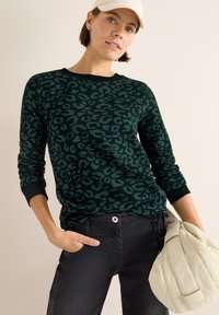 Felpa verde scuro con motivo leopardato, scollatura rotonda e polsini a costine. Abbinata a pantaloni neri e una borsa color crema.