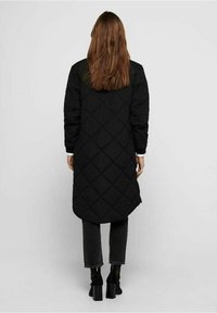 Manto lungo imbottito nero con orlo arrotondato, caratterizzato da maniche lunghe e un design semplice ed elegante. Indossato sopra jeans grigio scuro e stivaletti con tacco.