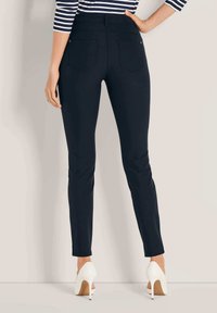 Vrouw gekleed in slim-fit zwarte broek en witte hoge hakken, staande met één hand op haar heup tegen een effen achtergrond.