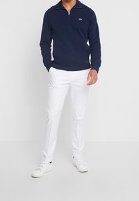 Mörkblå zip-up sweatshirt med ribbade muddar och nederkant, kombinerad med vita byxor och vita sneakers. Har en grön logotyp på bröstet.