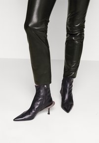 Zwarte leren enkellaarsjes met puntige neus, rits aan de zijkant en een unieke metalen accent op een stilettohak. Gecombineerd met glanzende groene broek.