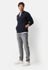 Navy zip-up sweatshirt with ribbed cuffs, paired with grey jeans and white sneakers. Model stands with hands in pockets against a plain background.