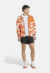 Orange og hvid mønstret jakke med lynlås foran, elastiske manchetter, båret over en hvid skjorte og sorte shorts, parret med hvide sportssko.