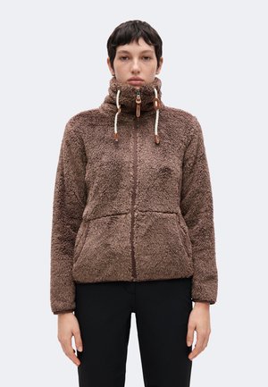 Persona que lleva una chaqueta de forro polar marrón con cremallera, cuello alto y cordones, de pie frente a un fondo claro y liso.
