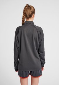 Haut de sport gris à manches longues avec un tissu texturé, col haut et manches raglan, associé à un short sombre avec des bordures rouges.