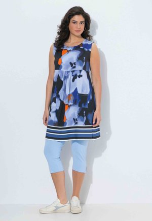 Mujer con cabello rizado que lleva una túnica sin mangas con un patrón en azul, negro y naranja, pantalones cortos azul claro y zapatillas blancas.