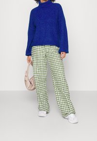 Blauer gestrickter Rollkragenpullover, lockere Passform. Weite Hosen mit grünem Hahnentritt-Muster. Hellrosa Tasche, weiße Sneakers.