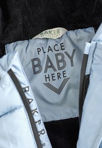 Heledad sinine pehmendus pehme musta karusnaha sisemusega. Tekst ütleb "TÕSTA BEBE SIIA" tugevas mustas kirjas. Silt ütleb "BAKER by Ted Baker."
