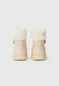 Bottines beige avec de gros revers en fourrure blanche pliés vers l’extérieur, vues de dos sur un fond clair uni.