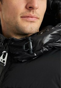 Detailný záber dolnej časti tváre a krku muža, ktorý má na sebe čiernu puffer bundu s viditeľným zipsom a šnúrkou na stiahnutie označenou "JACKIT."