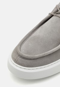Grå semsket slip-on sko med rund tå, polstret krage og hvit gummisåle med teksturerte detaljer og sømdetaljer.