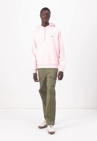 Sudadera con cremallera de color rosa claro con bolsillo tipo canguro, combinada con pantalones verdes oliva y zapatillas blancas con detalles en marrón. Diseño y corte simples.