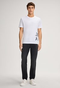 Weißes Baumwoll-T-Shirt mit Rundhalsausschnitt und grafischem Detail auf der linken Seite, kombiniert mit dunklen Slim-Fit-Jeans und weißen Sneakers.