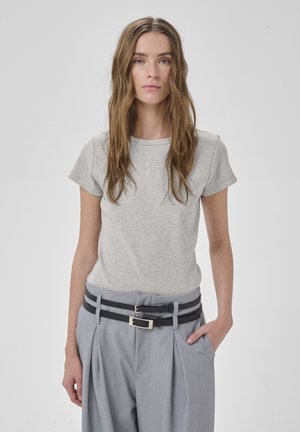 Noor pruunide pikkade juustega noor naine, kes kannab helehallit lühikeste varrukatega t-särki ja kõrge vöökohaga halli pükse kahe musta vööga.