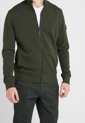 Mann trägt eine dunkelgrüne Jacke mit Reißverschluss über einem weißen Hemd und dunklen gemusterten Hosen, steht mit entspannter Haltung vor einem einfarbigen Hintergrund.
