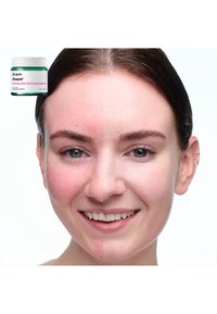 Dr. Jart+ Cicapair kremkrukke i grønn med hvitt lokk over et ansikt delt; venstre side viser rødhet, høyre side ser glatt og jevn ut.