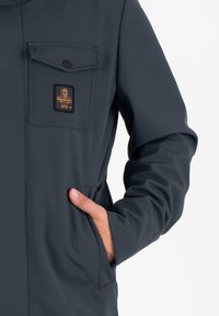 Donkergrijze jas met een borstzak aan de voorkant en een oranje logo patch. Gladde, matte textuur, aansluitend ontwerp met lange mouwen en zijzakken.