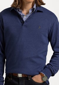 Tummansininen pitkähihainen poolopaita, jossa on kaulus, kaksi nappia ja pieni kirjailtu logo. Paita on pukeutunut raidallisen paidan päälle ja yhdistetty tummiin farkkuihin.