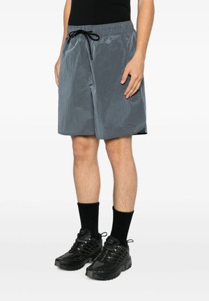 Uomo che indossa pantaloncini grigi alla lunghezza del ginocchio con coulisse nera, calzini neri e scarpe sportive nere, in piedi davanti a uno sfondo chiaro.