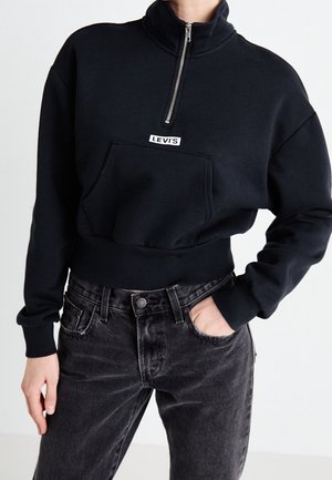 Person i en svart Levi's sweatshirt med kvart dragkedja och känguruficka, med en hand i fickan, mot en neutral bakgrund.