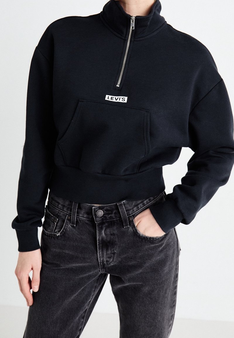 Persona che indossa un felpa nera Levi's a mezza zip con tasca a marsupio e jeans neri sbiaditi, con una mano in tasca, su uno sfondo neutro.